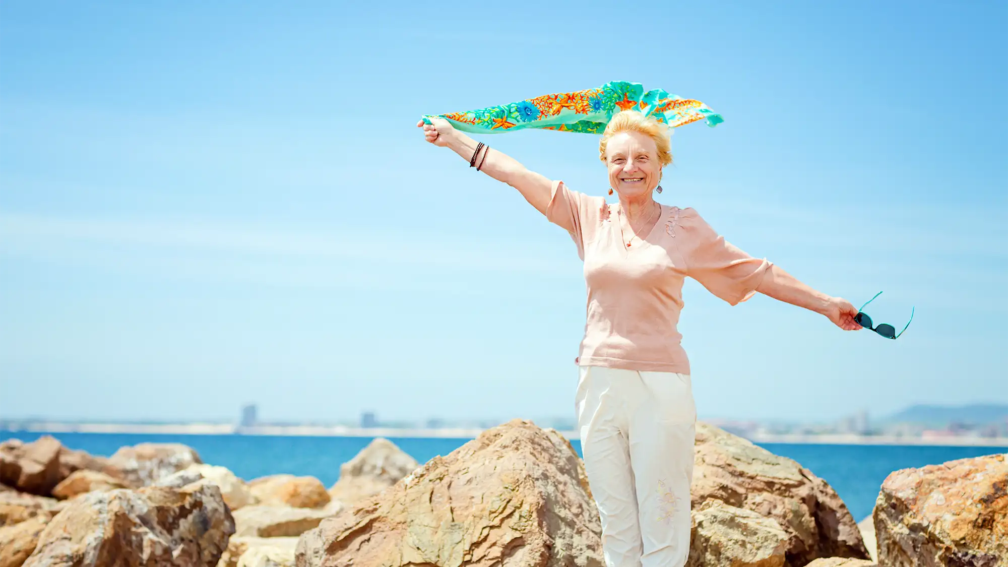 Mujer mayor sonriendo en unas rocas junto al mar, sosteniendo un pañuelo de colores al viento.