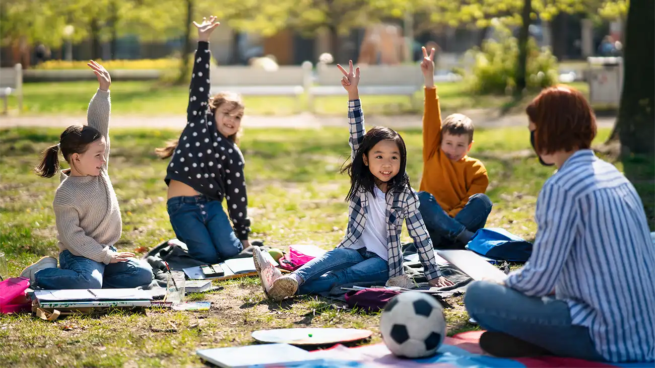 Un grupo de niños y una maestra en una clase al aire libre, sentados en el césped de un parque.