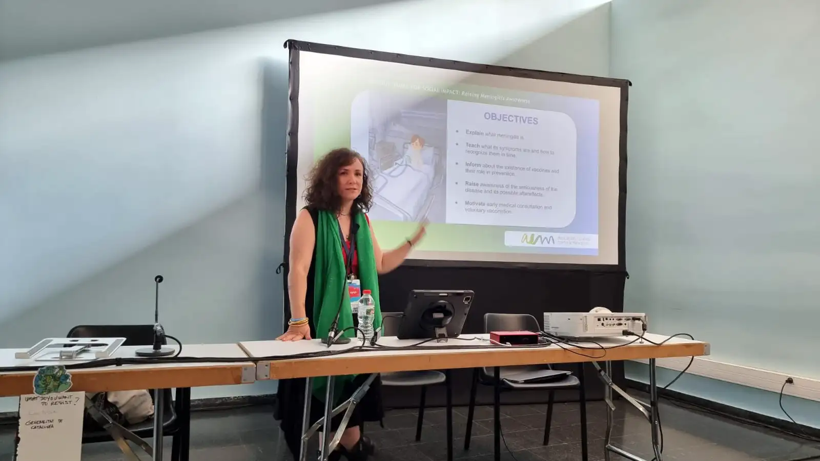 Una mujer dando una presentación sobre meningitis, de pie junto a una mesa y señalando una pantalla de proyector que muestra los 'Objetivos' y el logo de la AEM.
