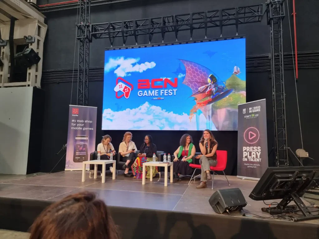 Un panel de cinco mujeres sentadas en un escenario durante el BCN Game Fest, con una gran pantalla del evento al fondo.