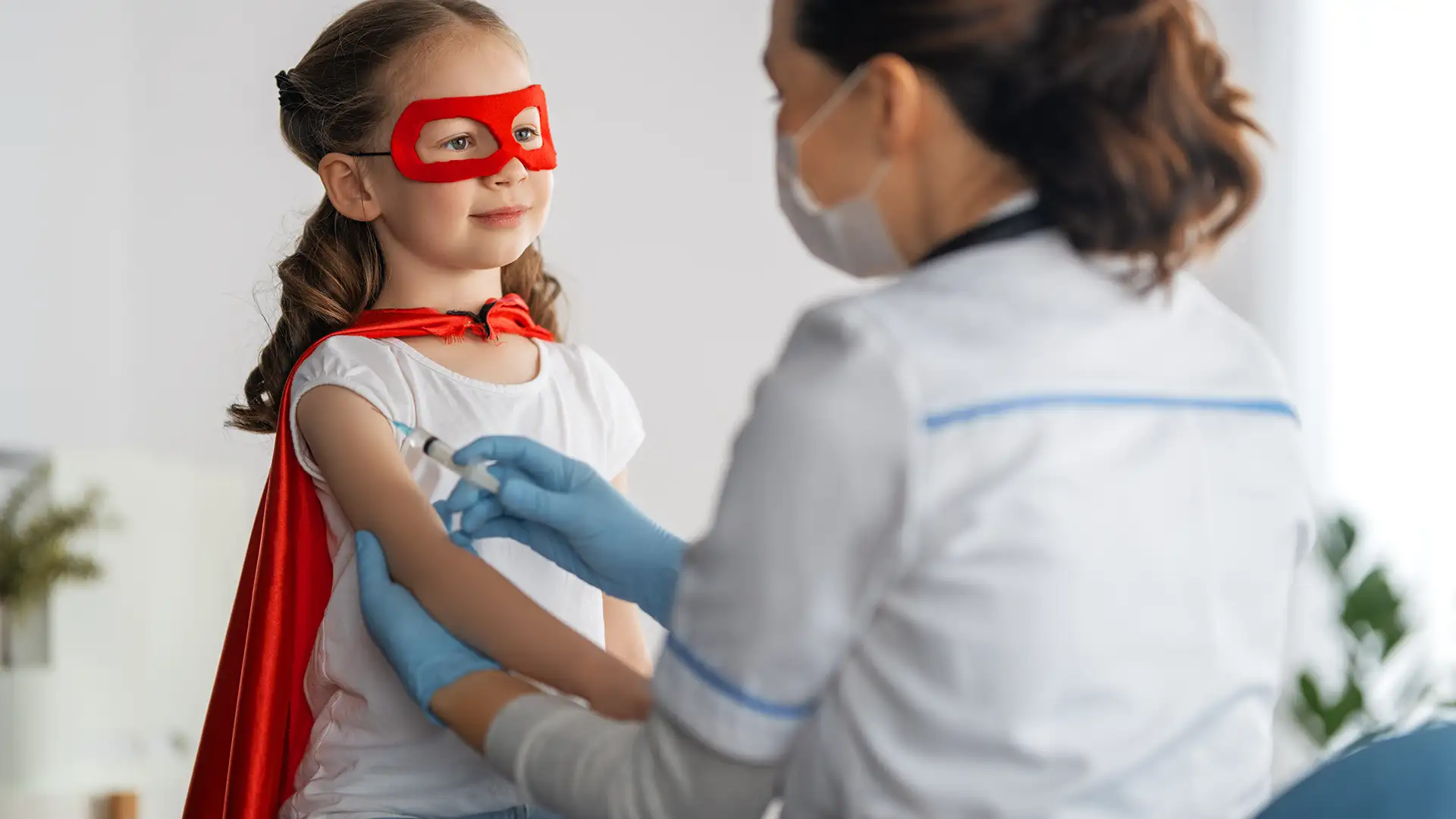 Una niña pequeña, disfrazada con una capa y un antifaz de superhéroe, recibe una vacuna en el brazo por parte de una profesional sanitaria.